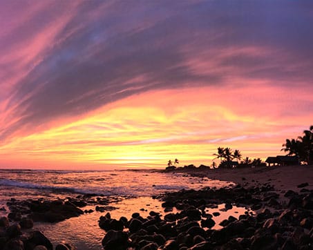Sun setting over a beach with gentle waves and mountainous terrain in the background under a dramatic sky.