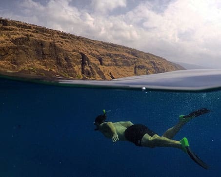 Snorkeler exploring underwater near a rocky coastline with a partly cloudy sky above.