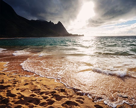 Sun setting over a beach with gentle waves and mountainous terrain in the background under a dramatic sky.